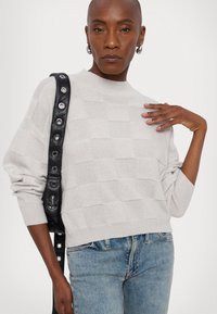 Maglione grigio chiaro, texturizzato, con un motivo a scacchi e colletto alto. Abbinato a jeans blu e una borsa a tracolla nera con dettagli in occhielli.