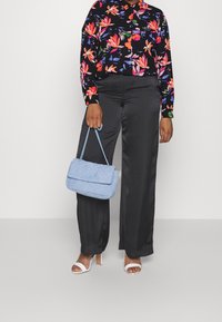 Personne portant une chemise à manches longues noire à fleurs, un pantalon large en satin noir, des sandales à talons blanches, tenant un sac à main matelassé bleu clair par ses poignées.