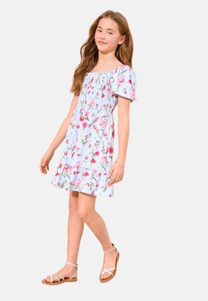 Mädchen mit langen Haaren, das ein blaues geblümtes Kleid und beige Sandalen trägt, steht mit einem leicht nach vorne gestellten Bein auf einem schlichten hellen Hintergrund.