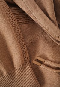Brązowy sweter z dzianiny, charakteryzujący się gładką strukturą, ściągaczami przy mankietach oraz kapturem, z dwoma beżowymi akcentami w postaci sznurków i starannie wykonanymi szwami.