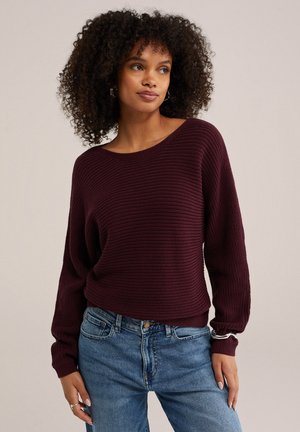 Kvinde med krøllet hår iført en mørkelilla strikket sweater og blå jeans, poserer med den ene hånd i lommen mod en ensfarvet baggrund.