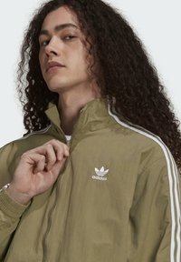 Person med långt lockigt hår som drar upp en olivgrön Adidas-jacka med vita ränder på ärmarna och logotyp på bröstet.