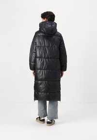 Abrigo acolchado largo negro con capucha, con un acabado brillante, diseño acolchado horizontal y longitud hasta la pantorrilla. Combinado con unos jeans holgados y zapatillas.