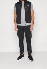 Czarna pikowana kamizelka na zamek z zielonym akcentem, noszona na szarym krótkim rękawie. Czarne spodnie typu jogger z kieszeniami na zamek i zielonymi detalami. Czarne sneakersy.