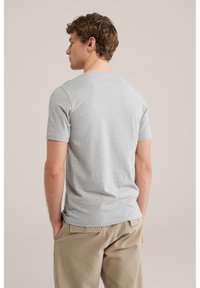 Muž otočený chrbtom, oblečený v svetlosivom tričku s krátkym rukávom a béžových nohaviciach, s rukami v vreckách, na jednoduchom pozadí.