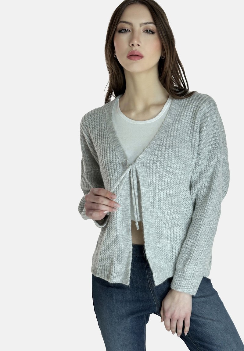 Cardigan lavorato a maglia grigio chiaro con design a V, dotato di chiusura con lacci sul davanti e spalle scese. Indossato sopra un top bianco.