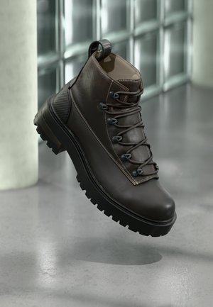 Bota de senderismo de cuero marrón con caña alta, suela de goma robusta, ojales metálicos y detalles cosidos. Cuenta con una lengüeta en la parte trasera.