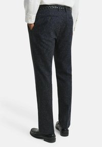 Mann in dunkel gesprenkelter Hose, schwarzem geflochtenem Gürtel, weißem Hemd und schwarzen Lederloafers, steht mit den Händen in den Taschen.
