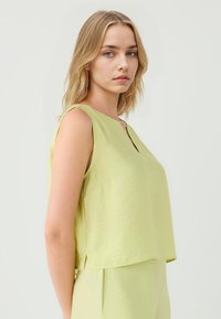 Giovane donna con lunghi capelli biondi posizionata in profilo laterale, indossa una maglietta senza maniche di colore verde chiaro su uno sfondo neutro.
