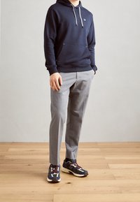 Sudadera navy con cordones, pantalones grises y zapatillas multicolores con un logo prominente y suela acolchada. Fondo de suelo de madera.