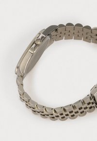 Hopeakylinen ruostumaton teräs kello, jossa on pyöreä kuori, uritettu kehys ja ketjuhihna, jossa on kaarevia ja litteitä osia.