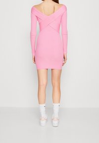 Donna che indossa un vestito aderente rosa a coste con schiena aperta incrociata, sneaker bianche Nike e calzini bianchi, in piedi su uno sfondo neutro.