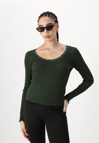 Mørkegrønn, ribbet langermet topp med en scallop-lace halslinje og uferdig kant, sammen med svarte tettsittende leggings og katteøyesolbriller.