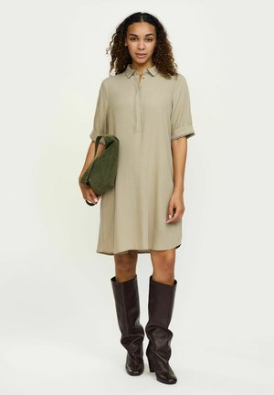 Vestido camisa caqui con cuello, mangas cortas abullonadas y detalles de botones. Llevado con botas altas de cuero marrón y sosteniendo un bolso verde.