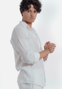 Witte linnen overhemd met lange mouwen, voorzien van een kraag en een ontspannen pasvorm. Het model staat met de handen gevouwen en kijkt iets naar links.