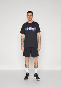 T-shirt preta de algodão com gráfico colorido "ADIDAS", combinado com calções pretos. Modelo usa ténis pretos e meias brancas até ao tornozelo.