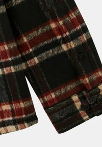 Chemise en laine à carreaux avec un motif à carreaux noir, rouge, beige et blanc, et boutons noirs sur les poignets et le rabat de poche.