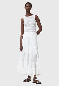 Abito maxi bianco a strati con un busto testurizzato e orlo arricciato. Il modello indossa sandali argentati, che mettono in risalto il design fluido dell'abito.