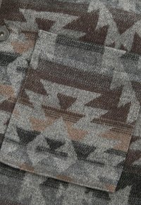 Vävt tyg med ett geometriskt mönster i nyanser av grått, svart och brunt. Inkluderar en ficka med lock och knappstängning.