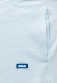 Ανοιχτό μπλε ύφασμα με μικρή μπλε ορθογώνια ετικέτα "HUGO" κοντά στη κάθετη ραφή της τσέπης στη δεξιά πλευρά.