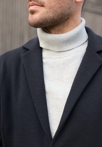 Man met lichtgrijze, geribbelde coltrui onder een donkerblauw colbert, getoond van de nek tot de bovenkant van de borst tegen een wazige achtergrond.