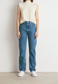 Cream sleeveless top, high-waisted blue jeans, and white sneakers. The jeans have a straight cut and classic five-pocket design. Soft texture.