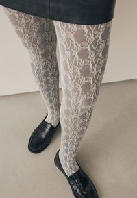 Collants blancs semi-transparents à motif de dentelle, associés à des mocassins noirs. Les jambes sont présentées sur un sol neutre.