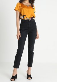 Žlutá saténová blůzka s květinovým potiskem, která je kombinována s vysokými černými džíny. Modelka má na sobě černé sandály na podpatku.