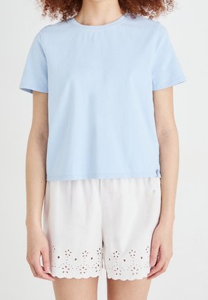 Persoon die een lichtblauw shirt met korte mouwen en witte korte broek met bloemige uitsnijdingen nabij de zoom draagt, staand tegen een effen achtergrond.