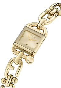 Zlato-tonirana ura s kvadratno številčnico, svetlo obarvano površino, zapestnico iz verižic in zaobljenimi detajli. Ima vgravirano blagovno znamko "Furla".