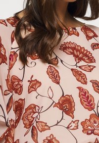 Kvinde iført en lys pink bluse med brune og orange blomster mønstre, viser bølget mørkebrunt hår og en del af halsen.