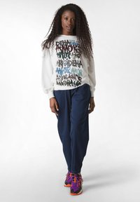 Sudadera blanca con texto gráfico colorido, pantalones azul marino y zapatillas multicolores. El modelo tiene el cabello rizado y está de pie.