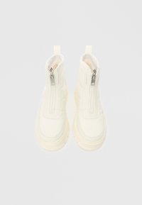 Et par hvite ankelstøvler med glidelås foran og tykke gummisåler, vist på en enkel lys bakgrunn.