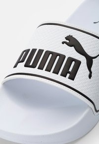 Biele šľapky vyrobené z textúrovanej gumy, s širokým popruhom s vyvýšeným čiernym logom „PUMA“ a siluetou čierneho mačka.