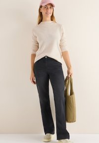 Beiger, gerippter Pullover mit langen Ärmeln, kombiniert mit dunkelblauen, geraden Hosen. Hält eine strukturierte, olivgrüne Tasche und trägt cremefarbene Sneakers.