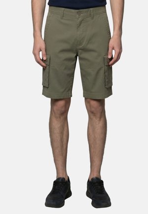Uomo con pantaloncini cargo verde oliva e scarpe da ginnastica nere, in piedi davanti a uno sfondo chiaro e uniforme.