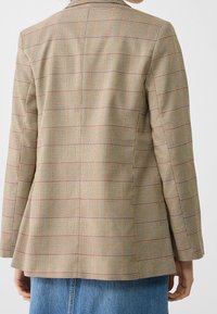 Persoon in een tan geruite blazer met rode en zwarte accenten, staande met ontspannen armen langs de zij, in combinatie met blauwe denim jeans.