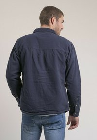 Chemise à manches longues bleu marine avec col, poignets boutonnés et détail de pli au dos. Fabriquée en tissu texturé. Portée avec un jean bleu clair.