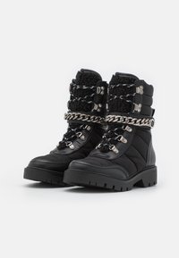 Botas de invierno negras con tejido acolchado, forro de borrego, ojales de metal, cordones en blanco y negro, y detalle de cadena plateada alrededor del tobillo.