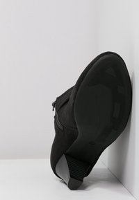 Botín de ante negro con tacón cuadrado apilado, cremallera lateral y suela de goma texturizada, inclinado contra una esquina blanca.