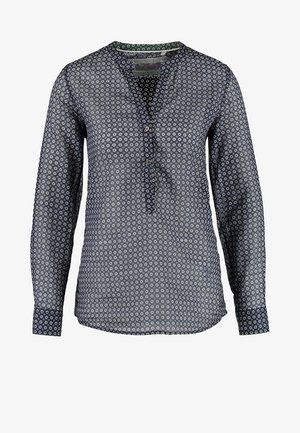 Frau trägt eine gemusterte Bluse, dunkle Skinny-Jeans, taupefarbene Stiefeletten und hält einen braunen breitkrempigen Hut in ihrer rechten Hand.