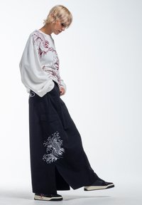 Hvid sweatshirt med røde fiskegrafikker, kombineret med sorte bukser med brede ben, der har et hvidt fiskedesign. Inkluderer sorte sneakers med røde detaljer.