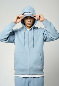 Sudadera con cremallera de color azul claro, con capucha ajustable, bolsillo tipo canguro y puños de canalé. Presenta un detalle de logo en el pecho y pantalones a juego.