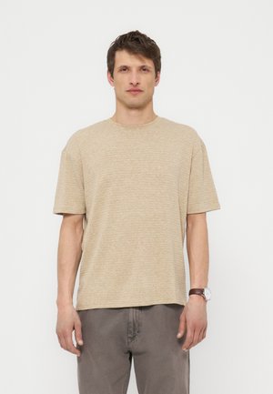 Mees, kes kannab beeži lühikeste varrukatega soonikutega t-särki ja halli pükse, seisab tavalise valge tausta ees, vasakul randmel käekell.