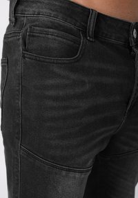 Μαύρο τζιν ύφασμα με λεία υφή, διαθέτει τυπικό κούμπωμα με κουμπί, μπροστινές τσέπες και διακριτικές ραφές.