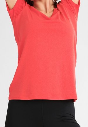 Vrouw draagt een koraalkleurig T-shirt met korte mouwen en V-hals en zwarte broek, met armen boven haar hoofd geheven tegen een effen achtergrond.