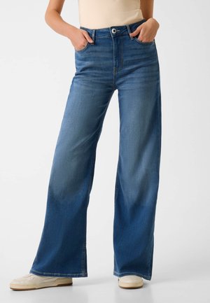 Kobieta nosząca niebieskie, szerokie dżinsy z wysokim stanem, z rękami w kieszeniach i białe trampki, stojąca na tle jednolitego jasnego tła.