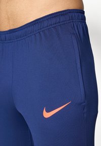 Dziļi zili sporta bikses no gludas auduma ar elastīgu jostu, sānu kabatām un oranžu Nike logo uz kreisās kājas.