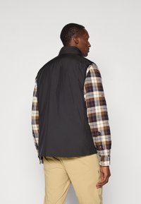 Black sleeveless vest made of smooth, lightweight material, worn over a plaid long-sleeve shirt with brown, blue, and cream colors.