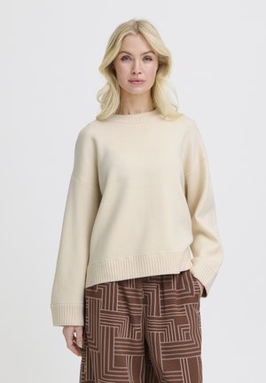 Femme aux cheveux blonds portant un pull crème et un pantalon large marron à motifs, debout devant un fond blanc uni.